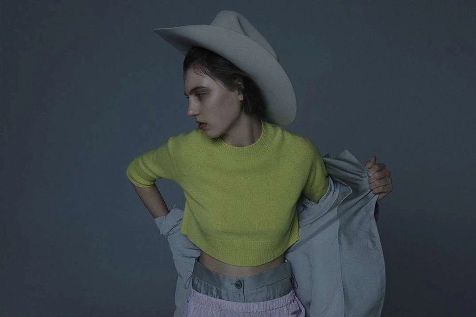 A fashion portrait of a young adult woman wearing a large hat, chartreuse top, and lilac pants in a photo studio setting.