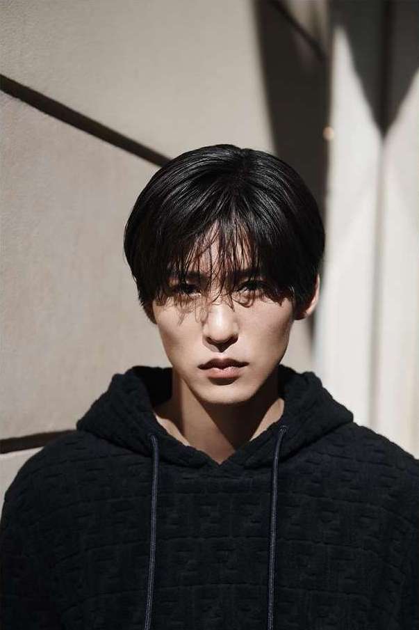 Close-up portrait of a young adult male with black hair and a textured black hoodie, posing against a beige neutral wall with shadow.