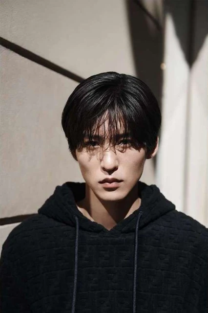 Close-up portrait of a young adult male with black hair and a textured black hoodie, posing against a beige neutral wall with shadow.
