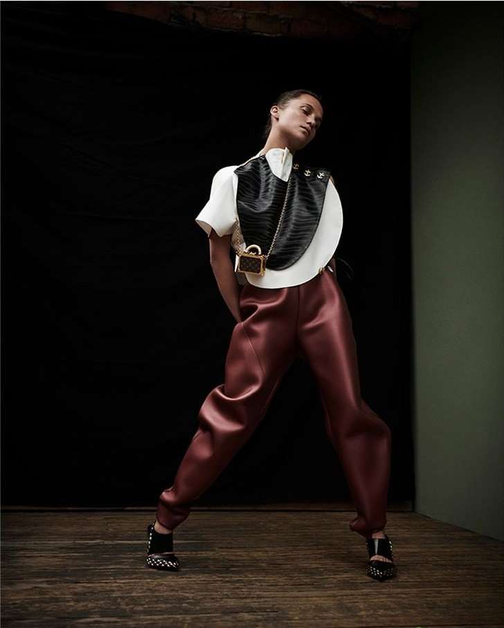 Woman in fashionable clothing poses in front of a black backdrop. She wears statement pants, a white top, and a black and white bag.
