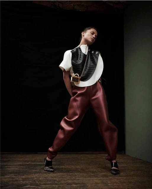Woman in fashionable clothing poses in front of a black backdrop. She wears statement pants, a white top, and a black and white bag.