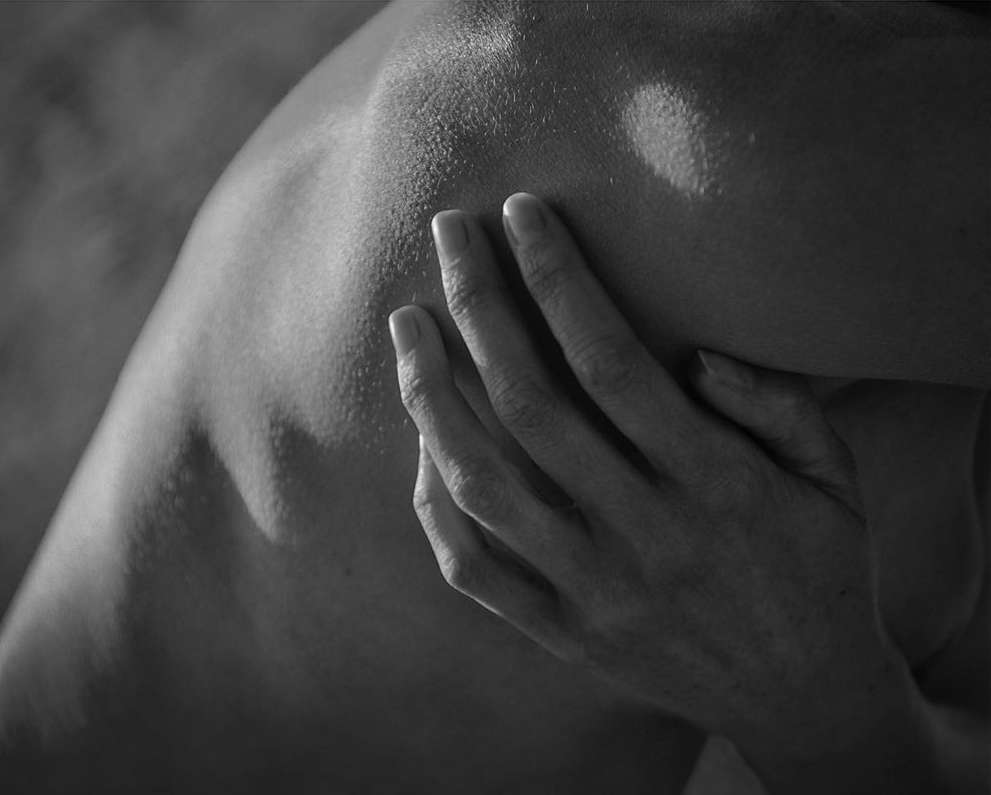 Monochrome studio portrait of an adult human's shoulder and hand, soft lighting creates a contemplative, intimate aesthetic.