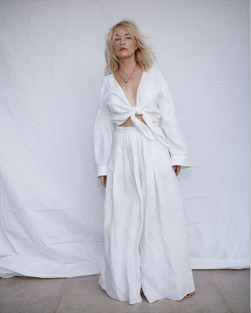 Woman in stylish white linen top and wide-leg pants against a minimalist backdrop, accessorized with layered necklaces, fashion shoot.