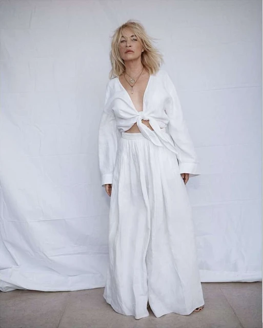 Woman in stylish white linen top and wide-leg pants against a minimalist backdrop, accessorized with layered necklaces, fashion shoot.