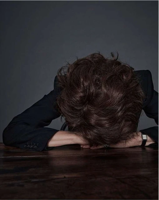 Person with curly brown hair in a dark suit leaning head-down on a dark wooden table against a dark grey background in a studio portrait.