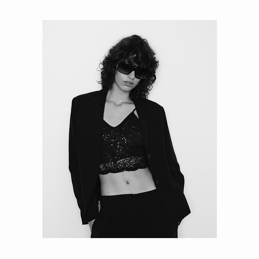 Black and white portrait of a woman with curly hair, wearing a black blazer, sequined top, and sunglasses against a white backdrop.