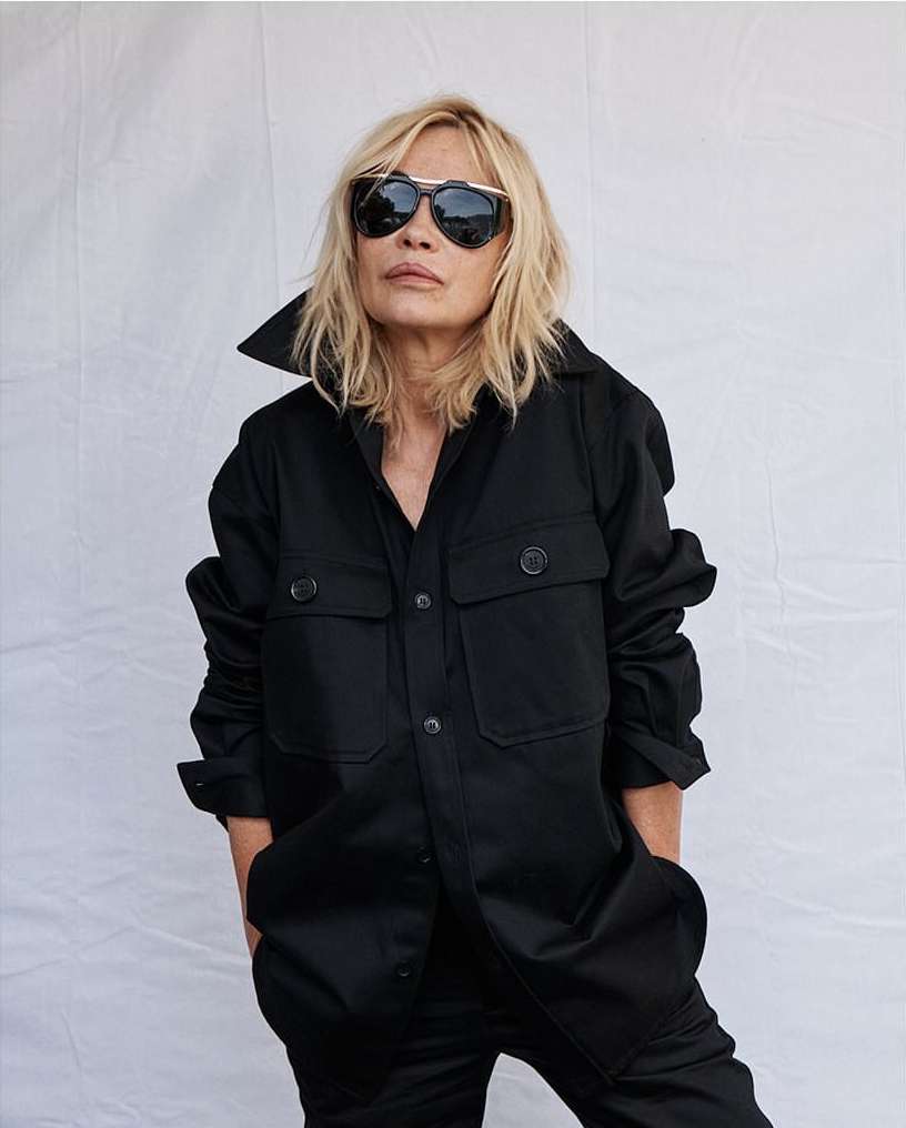 A blonde woman with sunglasses poses in a black jumpsuit against a neutral backdrop, showcasing a blend of casual and chic style.