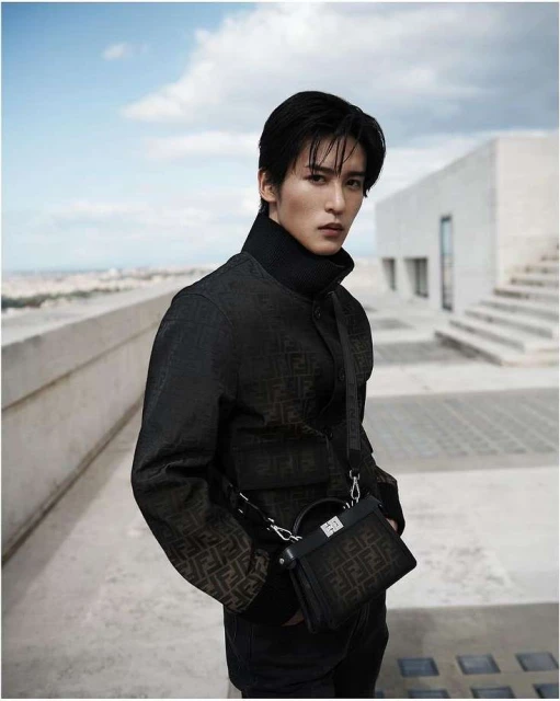 Man posing outdoors in Fendi jacket and bag, against modern architecture and blue sky. Fashion editorial shot, medium framing.