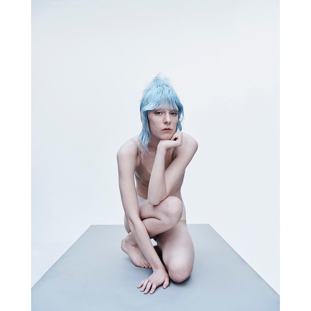 A portrait of an adult female with light blue hair, kneeling on a gray surface in a photo studio with a white background.
