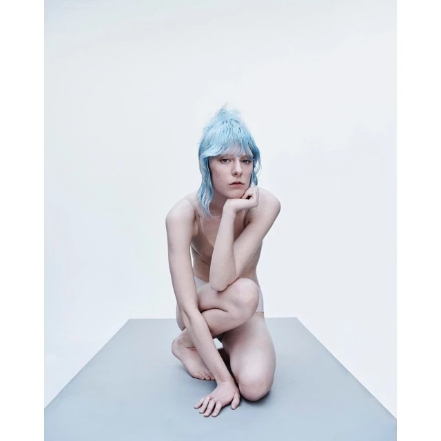 A portrait of an adult female with light blue hair, kneeling on a gray surface in a photo studio with a white background.