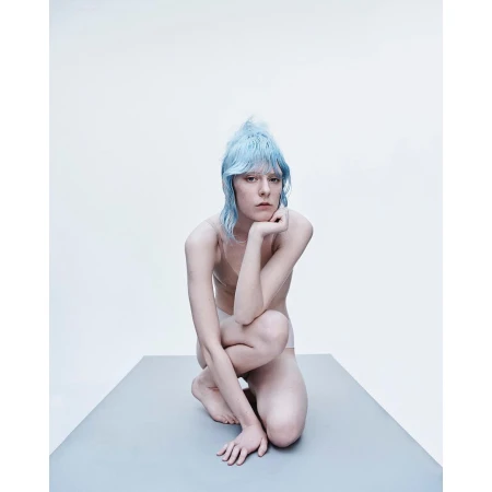 A portrait of an adult female with light blue hair, kneeling on a gray surface in a photo studio with a white background.