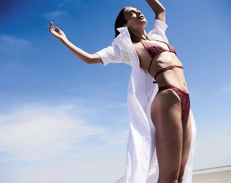 A woman poses in a printed bikini and a white cover-up against a blue sky at the beach on a sunny day.