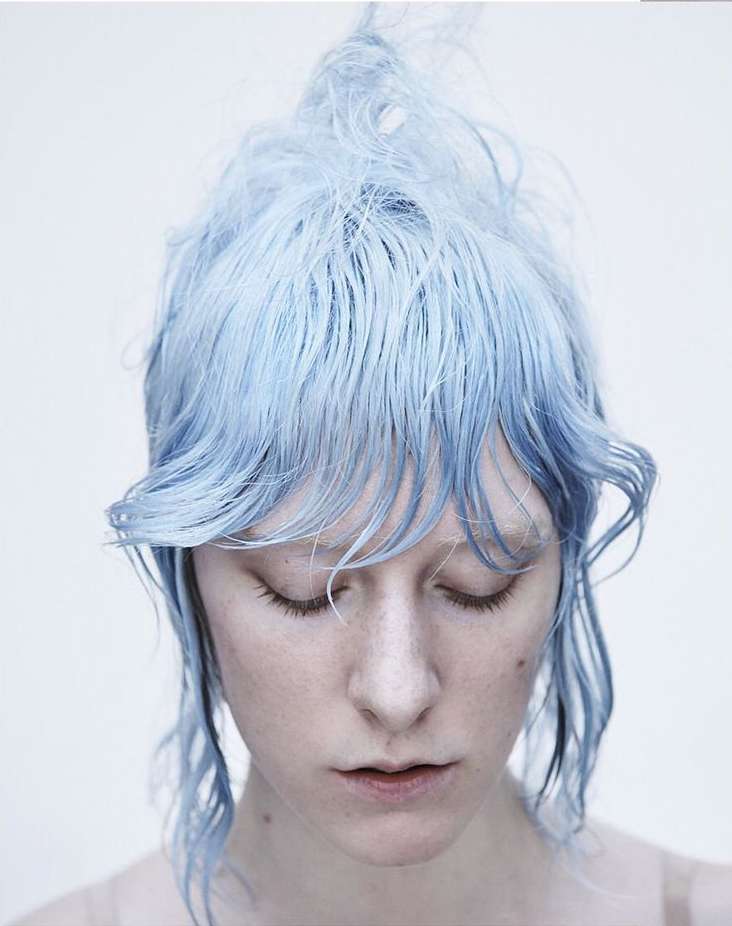 A portrait of a pale-skinned woman with closed eyes and striking light blue hair, against a bright, neutral background.