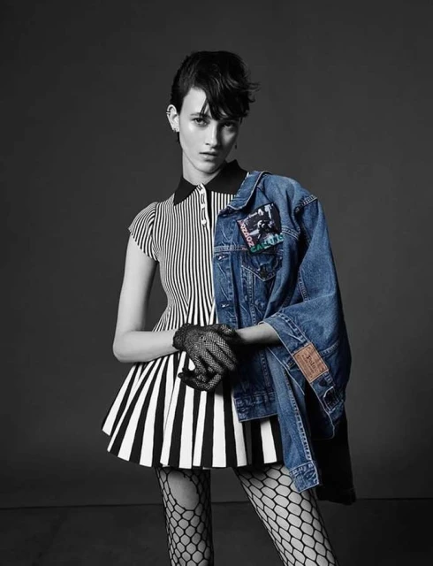A young adult fashion model poses in a black and white striped top and skirt with fishnet tights and a denim jacket on a gray backdrop.