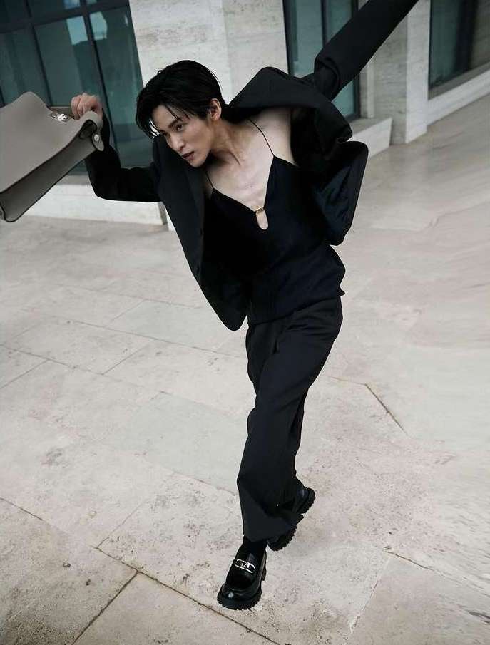 A male fashion model in a black suit strikes a dynamic pose outdoors holding a briefcase against a travertine stone building.