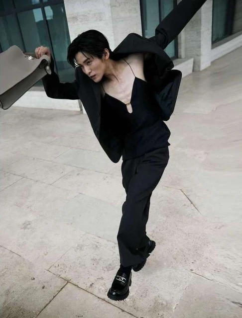 A male fashion model in a black suit strikes a dynamic pose outdoors holding a briefcase against a travertine stone building.