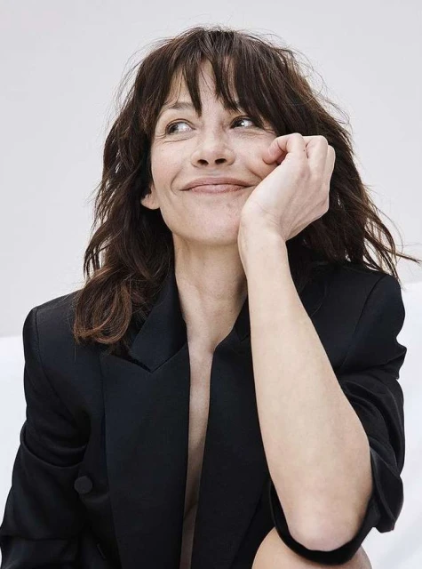 Close-up portrait of a brunette woman in a black blazer, hand to her face, set against a minimalist background.