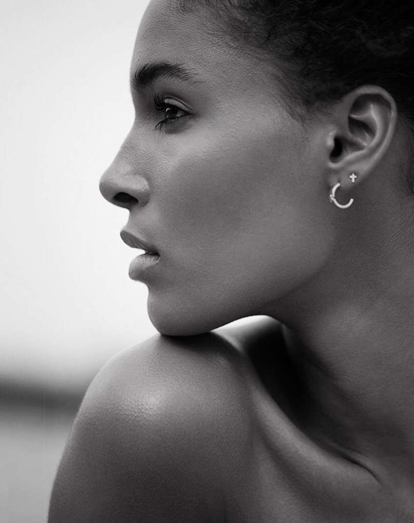 Close-up monochrome shot of a Black female adult facing left with bare shoulders, wearing hoop earrings against a blurred light background.