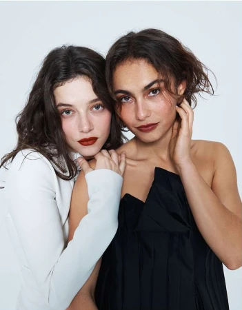 Two women pose together in a studio shot, one in a white jacket, the other in a black top, with dark lipstick and neutral eye makeup.