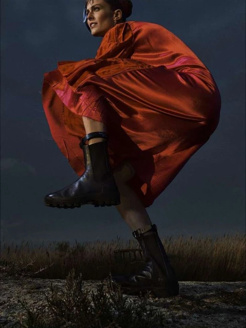 An adult female model in a red dress and black combat boots posing outdoors against a dusk sky in a stylized fashion shot.