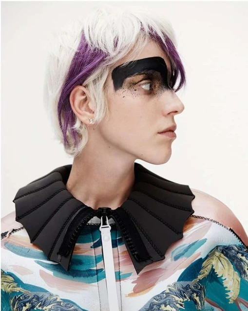 Profile of a woman with white and purple hair wearing black face paint and a jacket with a unique collar, captured in a studio shot.