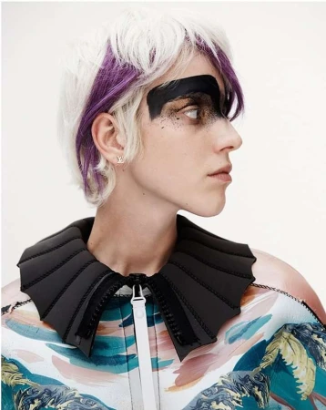 Profile of a woman with white and purple hair wearing black face paint and a jacket with a unique collar, captured in a studio shot.
