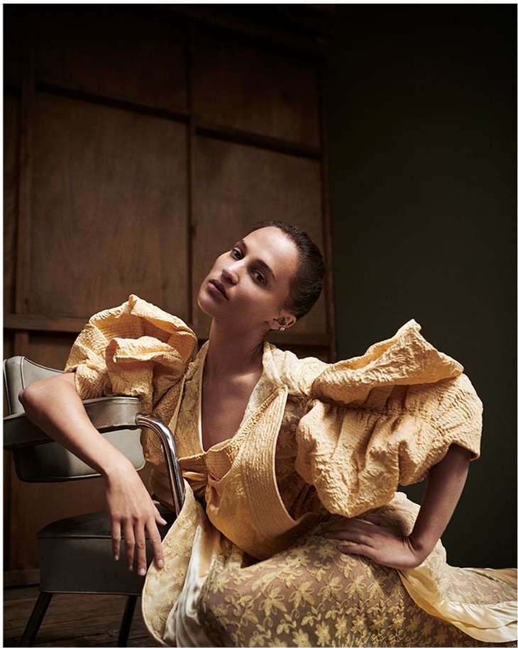 Indoor portrait of an adult female in a puffed-sleeve yellow dress, seated on a gray chair against a paneled brown wall.