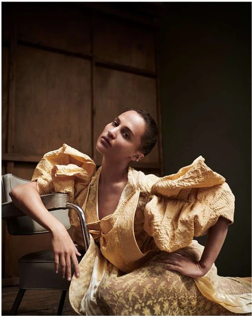 Indoor portrait of an adult female in a puffed-sleeve yellow dress, seated on a gray chair against a paneled brown wall.