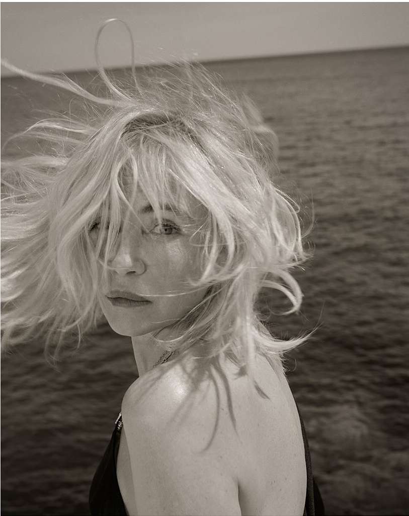 Monochrome portrait of a blonde woman with shoulder-length hair looking over her shoulder, with ocean water as a background.
