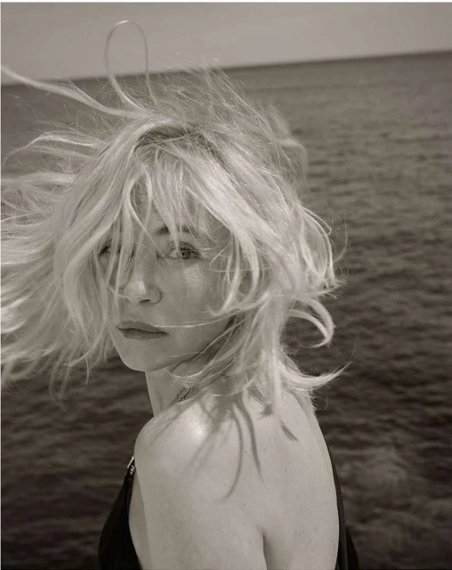 Monochrome portrait of a blonde woman with shoulder-length hair looking over her shoulder, with ocean water as a background.