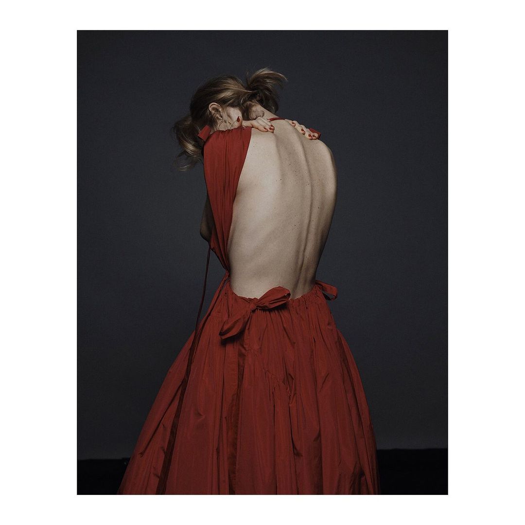 Back view of a woman in a red dress with a low back, her head bent forward, against a dark gray backdrop in a studio setting.