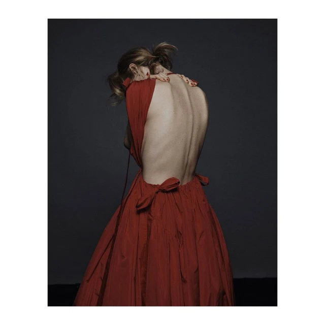 Back view of a woman in a red dress with a low back, her head bent forward, against a dark gray backdrop in a studio setting.