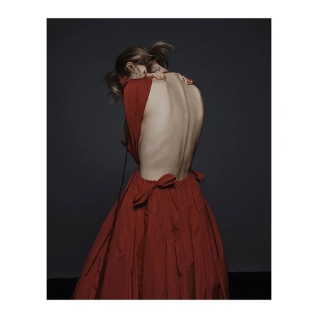 Back view of a woman in a red dress with a low back, her head bent forward, against a dark gray backdrop in a studio setting.
