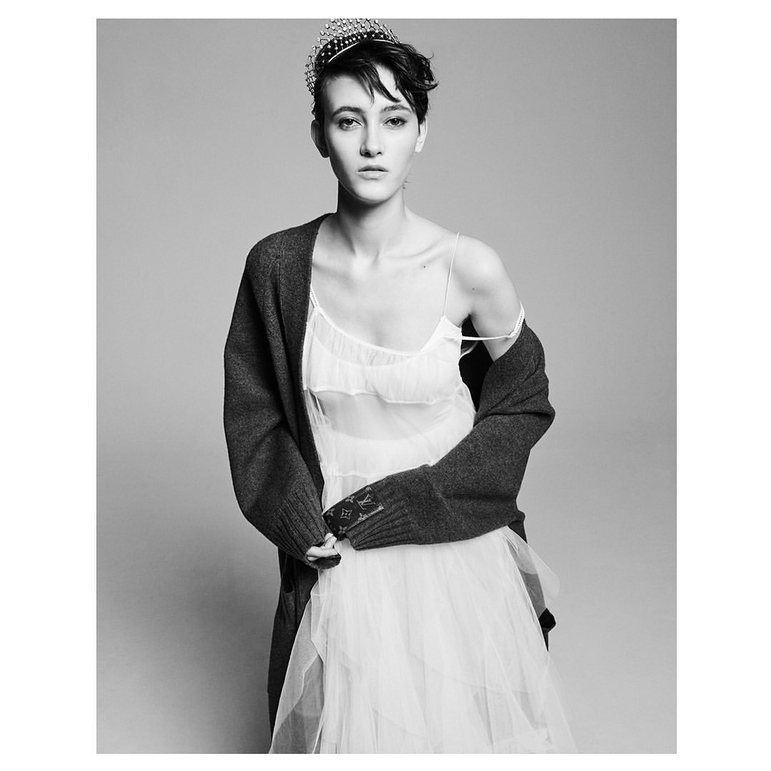 Black-and-white portrait of an adult woman wearing a crown, dress and cardigan, posing indoors in a photo studio against a grey backdrop.