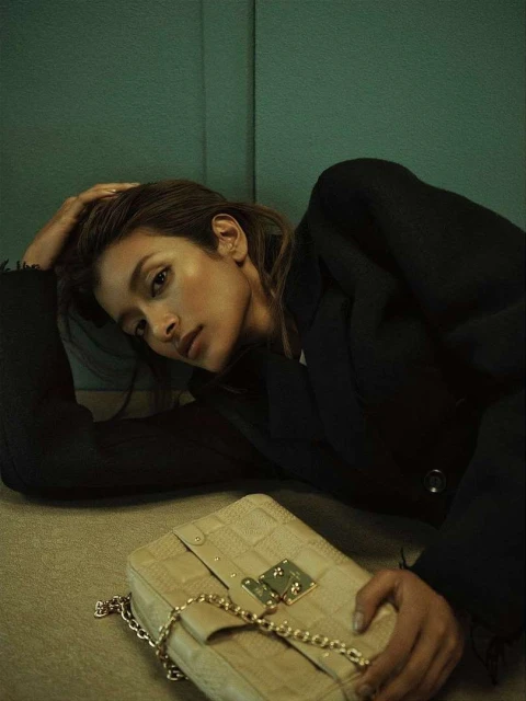 Woman poses with a light-beige handbag on a textured floor, wearing a black blazer, against a muted green wall in soft, diffused lighting.