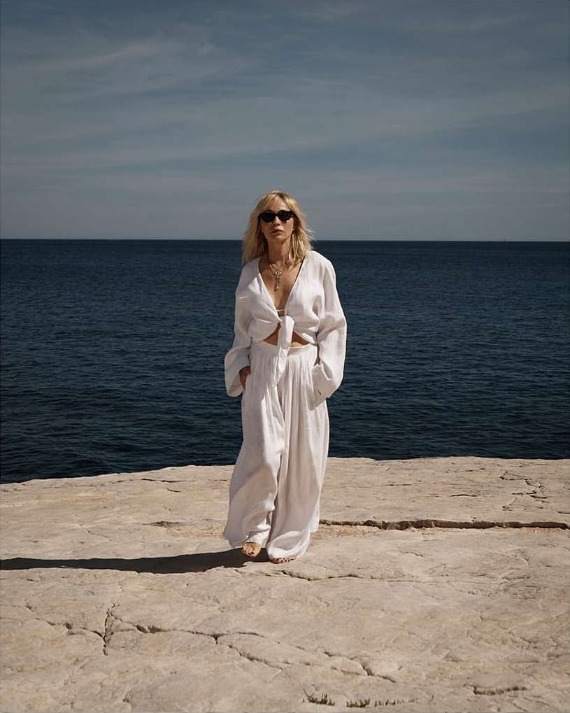 A blonde woman in a white linen outfit poses on a stone surface, with a blue ocean and sky in the background on a sunny day.