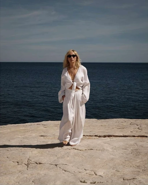 A blonde woman in a white linen outfit poses on a stone surface, with a blue ocean and sky in the background on a sunny day.