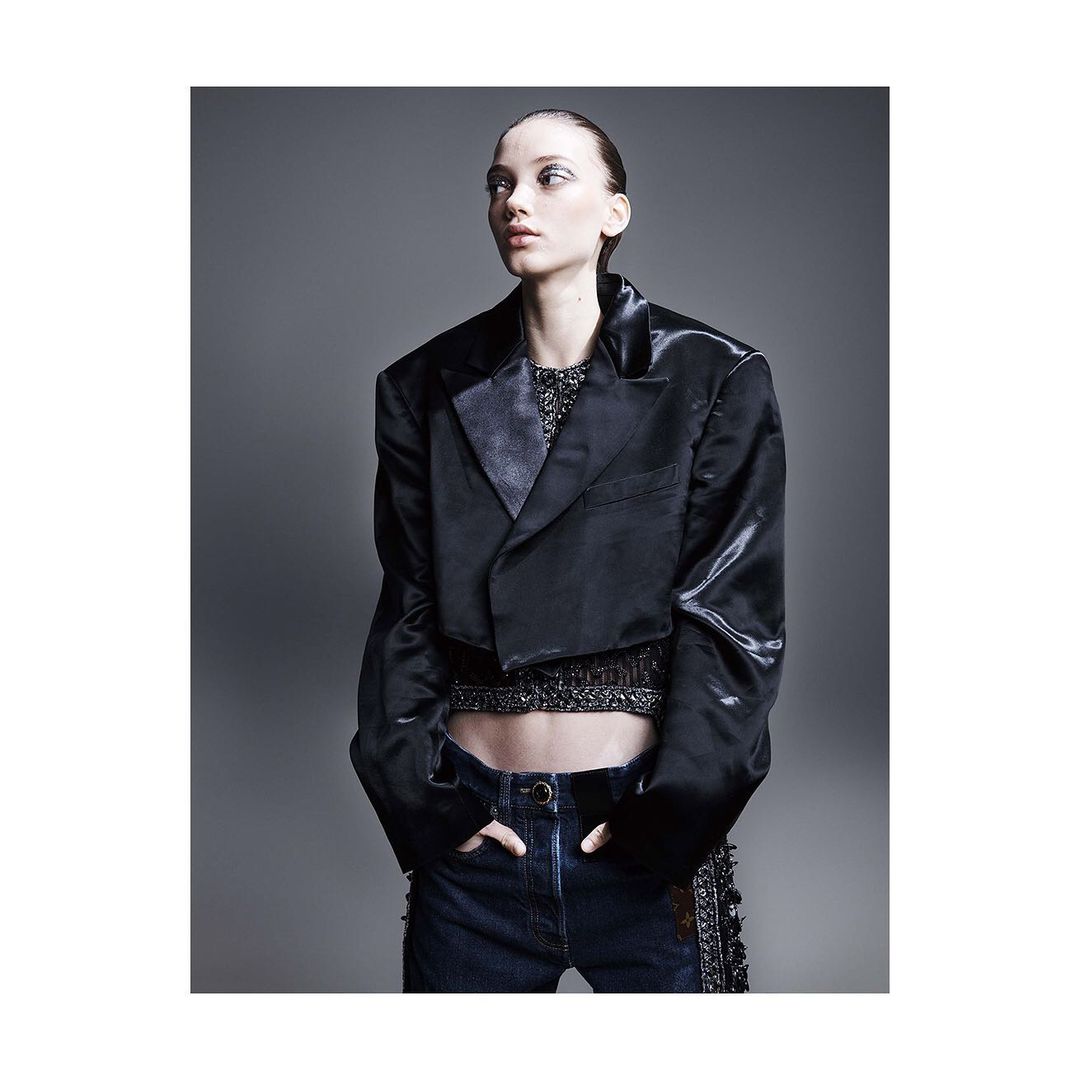 Fashion shot of a model in a dark jacket and jeans, posing in a photo studio with grey backdrop. The model is looking off to the side.