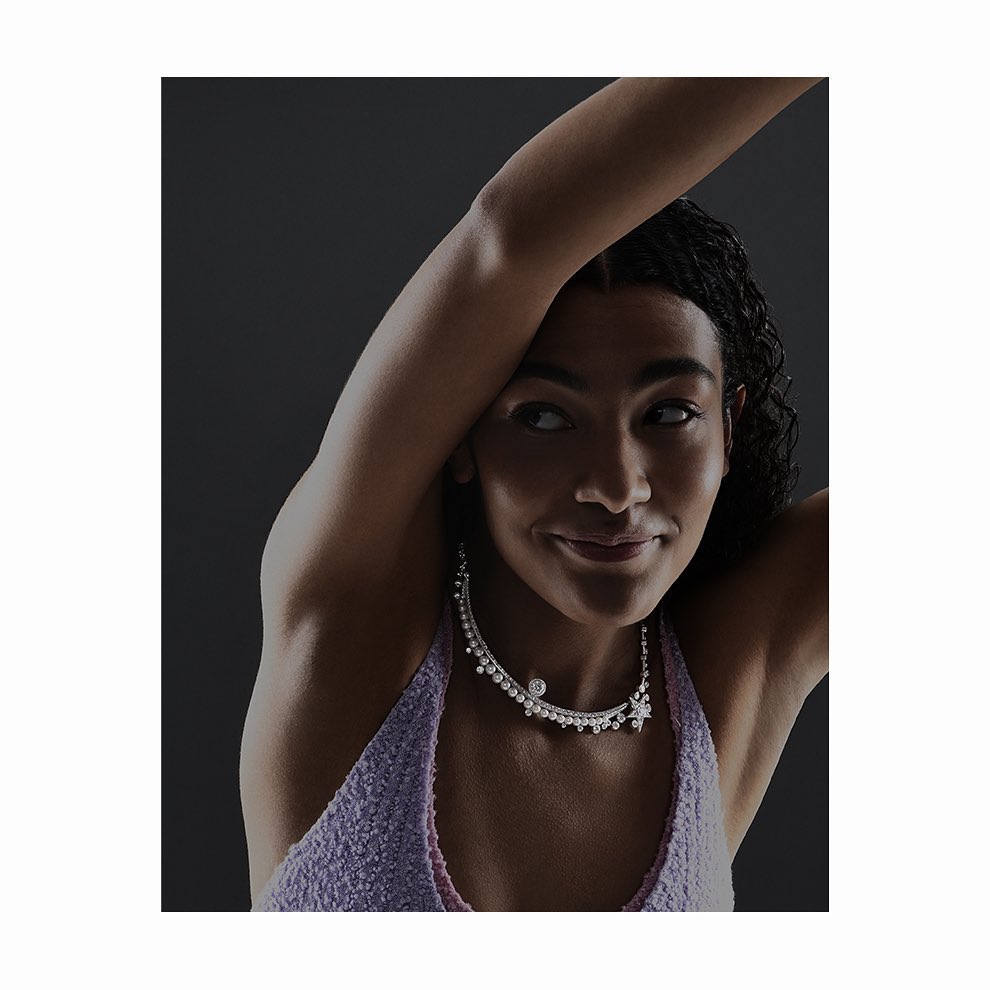 Elegant portrait of a young woman with arm raised, wearing a lavender top and embellished diamond necklace against a dark gray background.