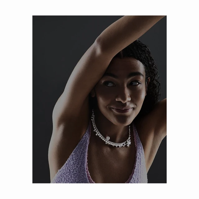 Elegant portrait of a young woman with arm raised, wearing a lavender top and embellished diamond necklace against a dark gray background.