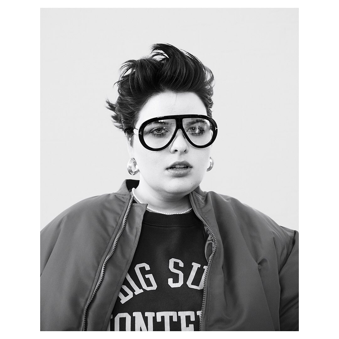 Monochrome portrait of a woman with short dark hair wearing aviator glasses, a bomber jacket, and a sweatshirt indoors against a light backdrop.