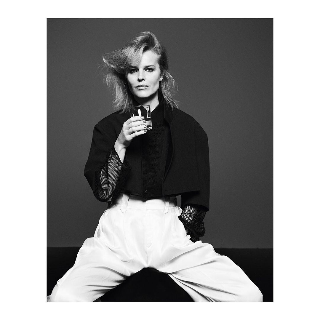 Black and white portrait of an adult female in a black blazer and white pants holding a glass, posing in a photo studio.