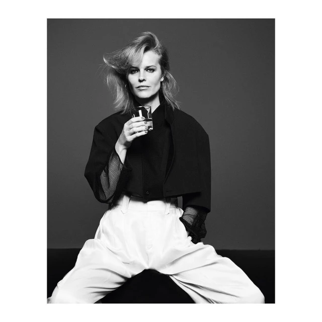 Black and white portrait of an adult female in a black blazer and white pants holding a glass, posing in a photo studio.