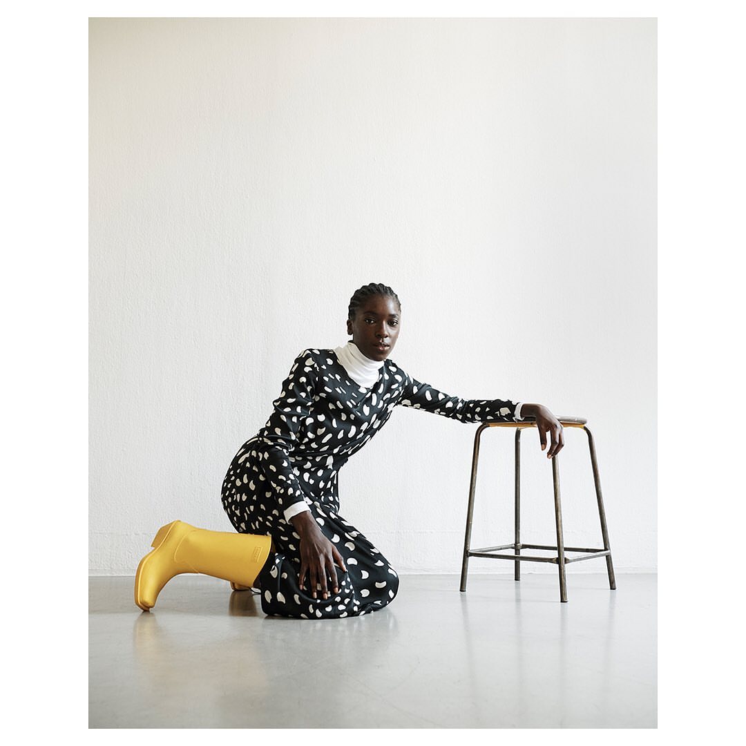 Fashion photography of a woman in a long-sleeved black and white dress, yellow boots, and kneeling next to a stool in a studio.