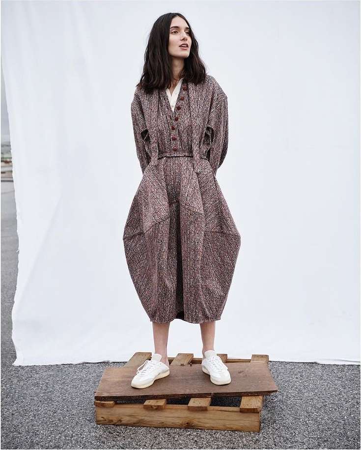 A slender woman with long, dark hair poses in a full-length shot wearing a unique brown dress and white sneakers while standing on a wooden pallet.