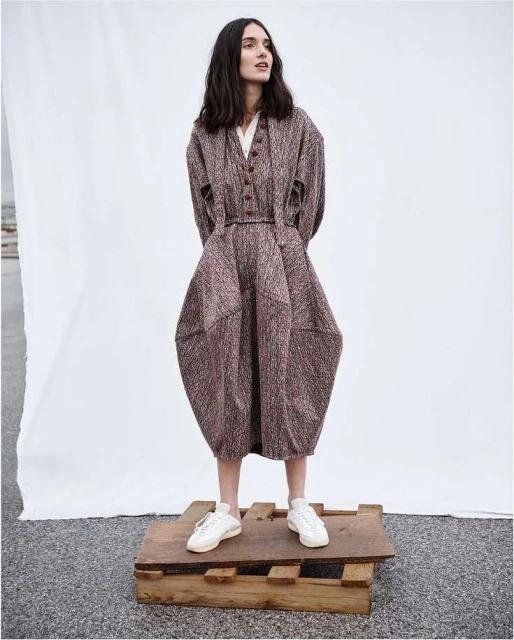 A slender woman with long, dark hair poses in a full-length shot wearing a unique brown dress and white sneakers while standing on a wooden pallet.