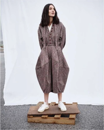 A slender woman with long, dark hair poses in a full-length shot wearing a unique brown dress and white sneakers while standing on a wooden pallet.