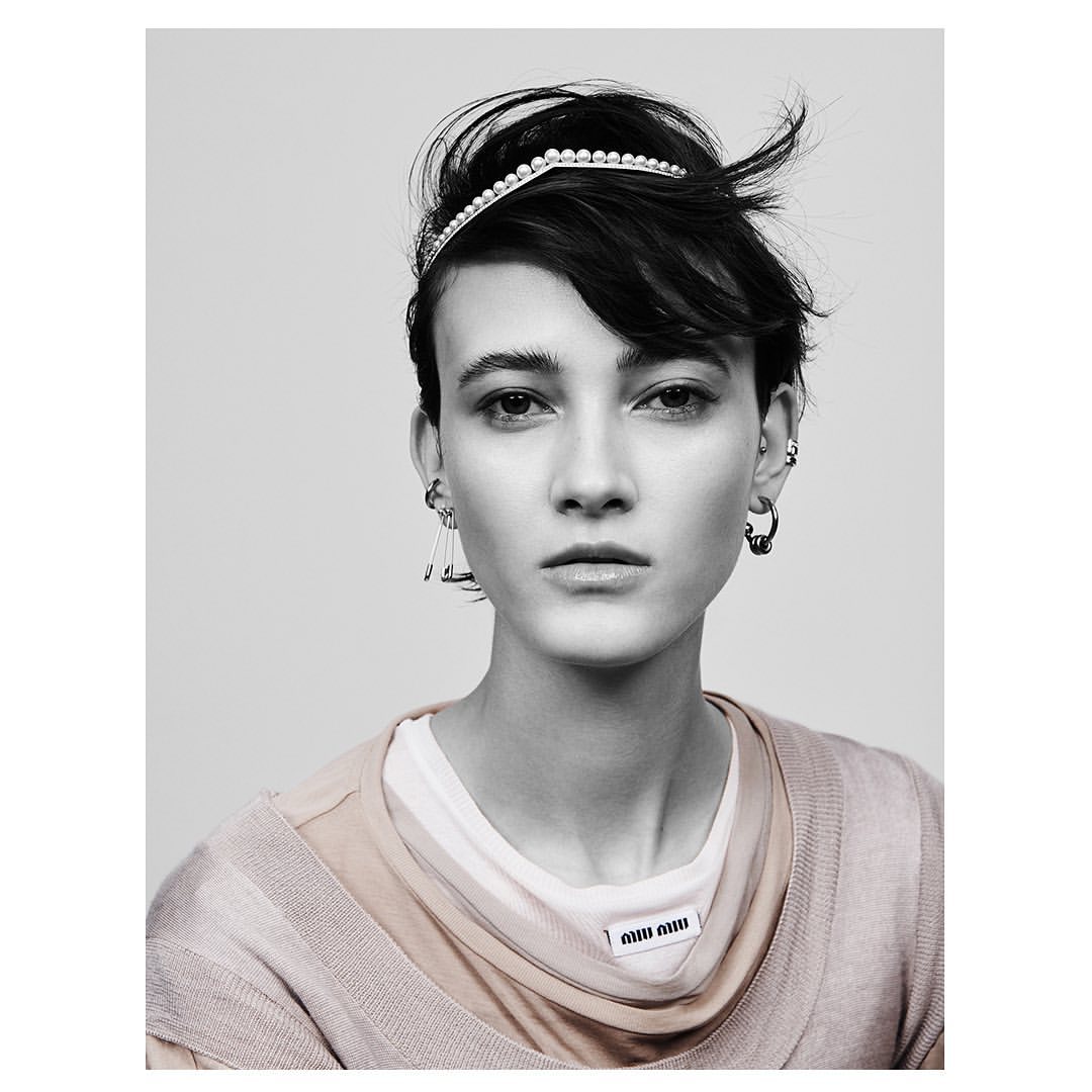 Close-up studio shot of a young adult female model with a pearl headband, neutral facial expression, and tan-colored layered top.