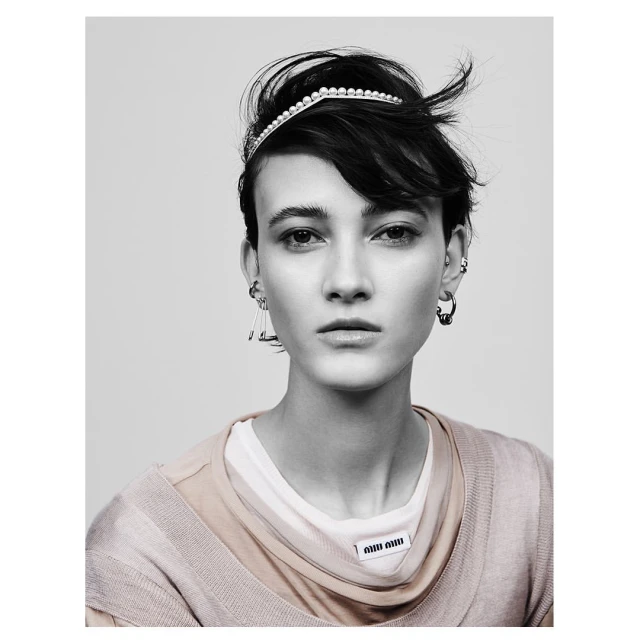 Close-up studio shot of a young adult female model with a pearl headband, neutral facial expression, and tan-colored layered top.