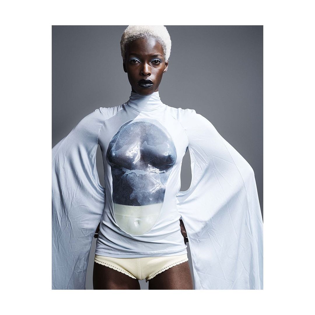 Striking studio portrait of a black female fashion model with white hair wearing a light blue long-sleeve top against gray backdrop.